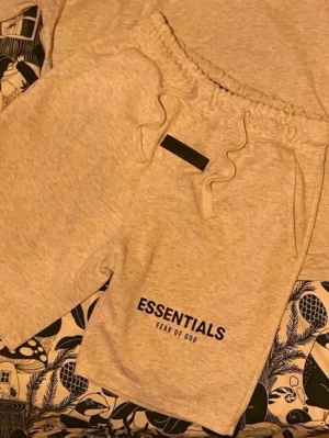 Grå shorts Essentials Fear of God - Gråa shorts från Essentials Fear of God med svart logga framtill. De har elastisk midja med snörning och är tillverkade i mjukt bomullsmaterial. Perfekta för en avslappnad och trendig stil. (Helt nya)-399 är minsta pris 