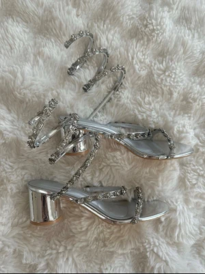 Silvriga sandaletter med strassdetaljer - Glansiga sandaletter i silver med blockklack och spiralformade remmar dekorerade med gnistrande strass. Öppen tå och trendig design som ger en riktigt statement-look. Perfekta för dig som vill sticka ut och addera lite extra bling till outfiten.