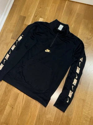 Svart Nike hoodie med dragkedja - Svart hoodie från Nike med halv dragkedja och stor ficka framtill. Guldiga Nike-loggor längs båda ärmarna och en på bröstet. Tillverkad i ett mjukt och sportigt material, perfekt för en avslappnad streetstyle-look.