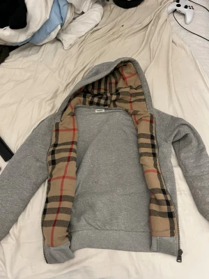 Grå burberry hoodie  - Snygg grå burberry hoodie med dragkedja och klassiskt beige, svart och rött rutigt foder i huvan och längs öppningen. Hoodien har en avslappnad passform och är perfekt för en chill och stilren look. Tillverkad i mjukt material för extra komfort.