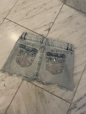 Ljus jeanskjol med rå kant - Säljer en kort ljusblå jeanskjol med fransig, rå kant och dekorativa bakfickor med broderi och knappdetaljer. Kjolen har bälteshällor och klassisk femficksdesign. Perfekt för dig som gillar en avslappnad och trendig stil.
