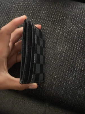 Svart rutmönstrad kortplånbok - Slimmad svart kortplånbok med stilrent rutmönster i mörkgrått och svart. Plånboken har flera kortfack och är tillverkad i ett material som ser ut som konstläder. Perfekt för dig som vill ha en enkel och snygg accessoar i fickan.
