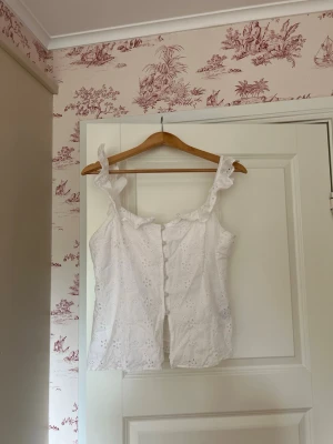 Vit broderad blus med volang - Söt vit blus med broderade detaljer och volangkant längs axelbanden. Blusen är ärmlös och har en feminin, romantisk look med knappar framtill. Perfekt till sommaren och passar till både jeans och kjol.