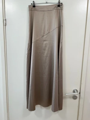 Beige långkjol i satin - Snygg beige långkjol i ett glansigt satinliknande material. Kjolen har en enkel, stilren design med en diagonal söm framtill som ger en cool detalj. Perfekt att styla med både sneakers och boots. Kjolen har en liten fläck därav har priset anpassats och har aldrigs används.