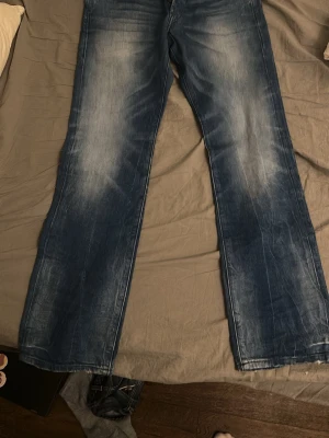 Blå bootcut jeans med slitningar - Säljer ett par blå bootcut jeans med snygga slitningar och tvättade detaljer på framsidan. Jeansen har klassisk femficksmodell och raka ben. Perfekta för en avslappnad och trendig look.
