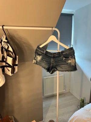 Blå slitna jeansshorts - Säljer ett par blå jeansshorts med slitningar och råa kanter. Shortsen har fem fickor, låg midja och klassisk knappgylf. Perfekta för varma dagar och ger en avslappnad look.