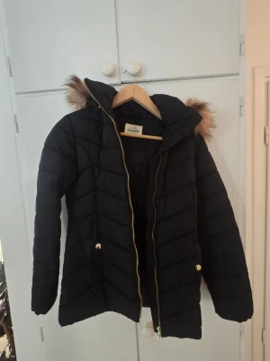 Svart dunjacka med fuskpäls KappAhl - Svart dunjacka från KappAhl med huva och fuskpälsdetalj. Jackan har guldfärgade dragkedjor och knappar, samt ett quiltat mönster. Perfekt för kalla vinterdagar och riktigt snygg till vardagslooken.