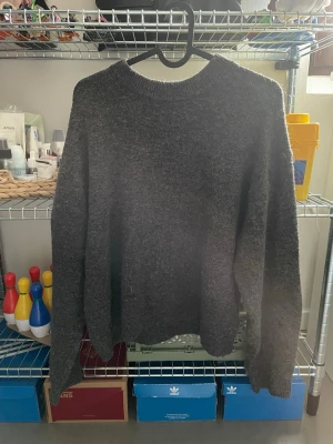Mörkgrå stickad tröja unisex - Mysig mörkgrå stickad tröja med rund hals och långa ärmar. Tröjan har en enkel, rak passform och är gjord i ett mjukt, fluffigt material som känns skönt mot huden. Perfekt för kyliga dagar och enkel att matcha med olika outfits.
