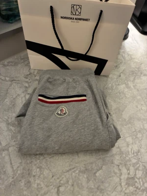 Grå shorts från Moncler med snörning - Snygga grå shorts från Moncler med snörning i midjan och fickor på sidorna. På ena benet finns Moncler-loggan och en rand i rött, vitt och blått. Perfekta för chill dagar eller när du vill ha en clean look. Nya pris 2000