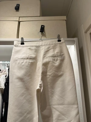 Vita raka kostymbyxor - Snygga vita kostymbyxor med raka ben och medelhög midja. Byxorna har två dekorativa bakfickor och är tillverkade i ett mjukt, vävt material som ger en clean och stilren look. Nästan helt oanvända. 32,5 cm i midjan, från skrevet till midjan 26 cm, innrebenslängd 66cm 