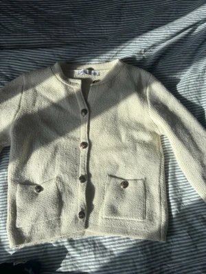 Krämvit kofta från Zara - Snygg krämvit kofta från Zara med rund halsringning och dekorativa knappar framtill. Koftan har två fickor fram och är stickad i ett mjukt material. Perfekt att slänga över en t-shirt eller linne för en chill look.