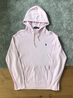 Ljusrosa hoodie från Polo Ralph Lauren - Ljusrosa hoodie med klassisk huva och vita dragsnören från Polo Ralph Lauren. Har en broderad marinblå logga på bröstet och en stor känguruficka framtill. Mjuk bomullskvalitet och avslappnad passform, perfekt för en chill look.