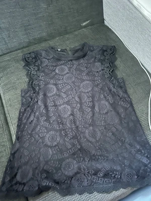 Svart spetsblus från Pieces - Säljer en svart blus från Pieces i storlek M. Blusen är ärmlös med vackra spetsdetaljer och ett genombrutet mönster över hela plagget. Perfekt för dig som gillar en feminin och trendig look med fina detaljer vid axlarna.