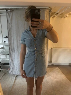 Kort Jeansklänning - Säljer en ljusblå kort jeansklänning med v-ringning och knappar framtill.  Den är avklippt där nere för att få till slitningar