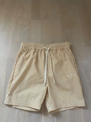 Randiga badshorts  från Les Deux - Säljer ett par gula och vita randiga badshorts från Les Deux. De har elastisk midja med snörning och sidofickor. Materialet känns lätt och luftigt, perfekt för varma dagar. Broderad logga på ena benet för en clean look.