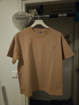 Beige t-shirt från Dior - Säljer en beige t-shirt från Dior med broderad logga på bröstet. T-shirten har rund halsringning och korta ärmar. Tillverkad i mjuk bomull och har en clean, stilren look som passar perfekt till jeans eller shorts.