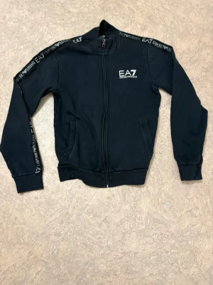 Svart zip hoodie från Emporio Armani - Snygg svart hoodie med dragkedja från EA7 Emporio Armani. Logga på bröstet och text längs ärmarna. Mjuk bomullskänsla, ribbade muddar och två fickor framtill. Perfekt för en sportig och trendig look.