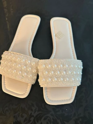 Vita sandaler med pärldekor 38  - Stilrena vita sandaler med fyrkantig tå och bred rem täckt av vita pärlor och små glittrande stenar. Snygg design med platt sula och mjukt material, perfekta för att lyfta din outfit med en elegant touch.