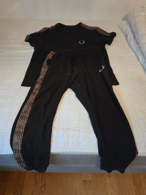 Svart loungewear-set med guldiga detaljer - Mysigt svart loungewear-set med t-shirt och byxor. Setet har guldiga ränder längs axlar och byxben samt små guldfärgade emblem på bröstet och byxan. Byxorna har dragsko i midjan och resår vid bensluten. Perfekt för chill dagar hemma. Storlek m