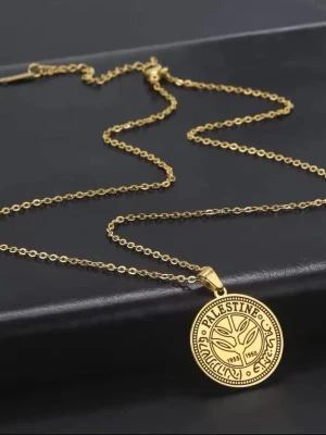Guldhalsband med Palestina-myntberlock - Snyggt halsband i guldfärgad metall med en rund berlock som liknar ett gammalt palestinskt mynt. Berlocken har detaljerade mönster och texten 'Palestine' samt årtalen 1955 och 1950. Kedjan är tunn och elegant, perfekt för att ge din outfit en unik touch.