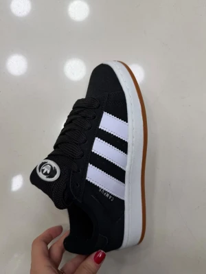Svarta sneakers med vita ränder , storlek 37 - Snygga svarta sneakers från Campus med klassiska vita ränder på sidorna och brun sula. Skon har svart ovandel i textil och syntet, vita detaljer och svart snörning. Perfekt för en sportig och stilren look.