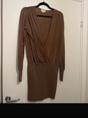 Omlottklänning - Snygg kort brun klänning med glittrigt tyg och omlottmodell. Djup v-ringning och långa ärmar ger en trendig look. En perfekt fest eller nyårklänning  som sitter galet snyggt på en.  Har dock en reva vid midjan, men är enkelt att sy ihop.