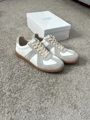Maison Margiela vita och grå sneakers - Snygga sneakers från Maison Margiela i vitt och ljusgrått läder och mocka. Skorna har beige snören och en klassisk gummisula i brunt. Perfekta för dig som gillar stilrena och trendiga detaljer. Kommer med originalkartong.