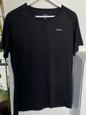 Svart tränings-tshirt från Craft XL  - En stilren svart tränings-tshirt från Craft med rund halsringning och korta ärmar. Tillverkad i ett lätt och snabbtorkande funktionsmaterial som passar perfekt till löpning eller gym. Diskret logga på bröstet.
