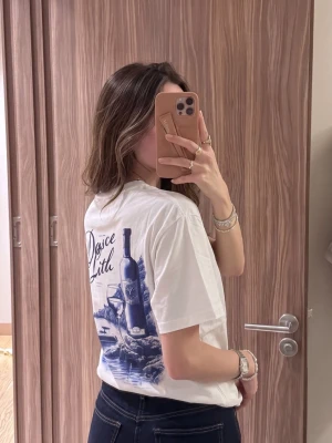 Vit t-shirt med blått vintema-tryck - Säljer en vit t-shirt med ett stort blått tryck på ryggen som föreställer en vinflaska, två glas och texten 'Dolce Litu'. T-shirten har avslappnad passform och korta ärmar. Perfekt för dig som gillar unika motiv och vill sticka ut lite extra.