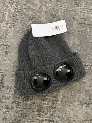 Grå ribbstickad mössa med glasögondetalj - Snygg grå ribbstickad mössa med uppvikt kant och två svarta glasögonliknande detaljer framtill. Mössan har en modern streetwear-vibe och är tillverkad i mjukt stickat material. Perfekt för dig som vill sticka ut med din stil.
