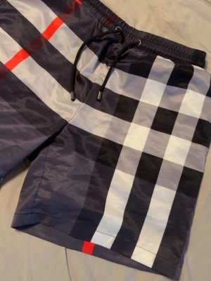 Rutiga badbyxor med dragsko - Snygga badbyxor med klassiskt rutmönster i svart, vitt och grått samt röda detaljer. De har elastisk midja med svart dragsko och en bakficka. Materialet är glansigt och snabbtorkande, perfekt för stranden eller poolen.