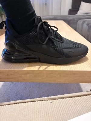 Svarta Nike Air Max 270 sneakers - Säljer ett par helsvarta Nike Air Max 270 sneakers med mesh-ovandel och stor synlig Air-enhet i hälen. Skorna har en sportig look med diskreta loggor och snörning. Perfekta för dig som gillar stilrena och bekväma sneakers.