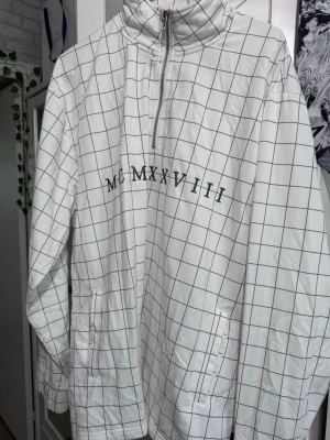 Vit rutig polotopp från Topman - Säljer en vit polotopp från Topman med svart rutmönster och trycket MCMLXXVIII på bröstet. Toppen har half zip-dragkedja, hög krage och två fickor framtill. Perfekt för dig som gillar stilrena och moderna plagg.