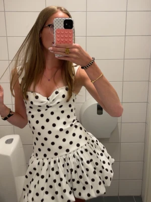 Vit ny miniklänning med svarta prickar - Supersöt vit miniklänning med svarta prickar och breda axelband. Klänningen har en rynkad byst, figurnära midja och voluminös kjol med ballongform. Perfekt för dig som gillar retrovibbar och lekfull stil.