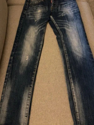 Blå dsquared jeans  - Säljer ett par blå jeans med slitningar och ljusa partier framtill och baktill. Jeansen har raka ben och tryckt text på baksidan. Klassisk femficksmodell i denim med coolt sliten look.28w 32L