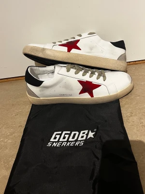 Vita GGDB sneakers med röd stjärna - Snygga vita sneakers från Golden Goose med en ikonisk röd stjärna på sidan, beige sula och grå skosnören. Hälpartiet är svart och skon har en klassisk låg profil. Materialet är läder och sulan är i gummi. Perfekta för en trendig och avslappnad stil.