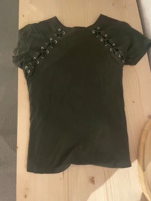 Olivgrön t-shirt med snörning - Snygg olivgrön t-shirt med cool snörningsdetalj över axlarna. T-shirten har korta ärmar. Perfekt nu till hösten. 