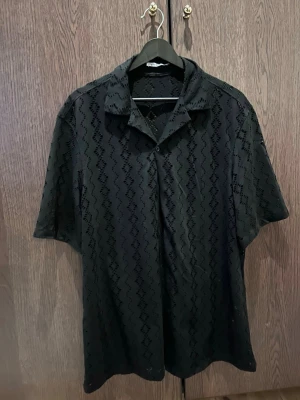 Svart meshskjorta med mönster - Säljer en svart skjorta från Zara i meshmaterial med vågigt hålmönster. Skjortan har korta ärmar, klassisk krage och knäppning framtill. Perfekt för dig som vill ha en luftig och trendig look.