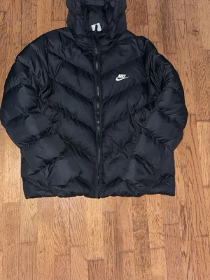 Svart Nike pufferjacka Therma-FIT XL - Säljer en svart pufferjacka från Nike med Therma-FIT-teknologi. Jackan har huva, dragkedja framtill och Nike-logga på bröstet. Fylld för extra värme och perfekt för kalla dagar. Passar dig som vill ha en stilren och sportig look.