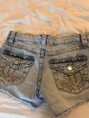 Ljusblå  jeansshorts med paljetter - Säljer ett par ljusblå jeansshorts med håliga detaljer. Shortsen har två bakfickor med broderade mönster och paljetter för extra bling. Klassisk femficksmodell med bälteshällor och knappstängning framtill.