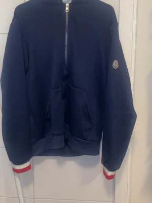 Mörkblå hoodie med dragkedja - Snygg mörkblå hoodie med hel dragkedja framtill och fickor. Har Moncler-logga på ärmen och ribbade muddar med röd och vit rand. Klassisk huva och sportig look, perfekt till streetwear eller chill dagar.