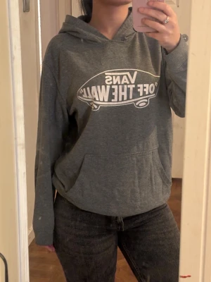 Grå hoodie från Vans med tryck - Säljer en grå hoodie från Vans med det klassiska 'Off The Wall'-trycket i vitt framtill. Hoodien har huva, magficka och ribbade muddar vid ärmslut och nederkant. Perfekt för en avslappnad och cool stil. Xl barn så passar xs/s