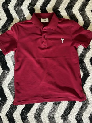 Ami polo - Snygg vinröd pikétröja med klassisk krage och knäppning framtill. På bröstet finns en vit broderad detalj med hjärta och bokstaven A. Tröjan är i ett mjukt bomullstyg och har korta ärmar, perfekt för en avslappnad men stilren look.