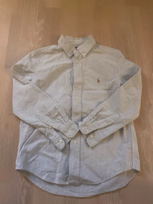 Randig skjorta med broderad logga - Klassisk skjorta med blå och vita ränder och broderad logga på bröstet. Skjortan har knappar framtill, button-down-krage och långa ärmar med manschett. Tillverkad i ett mjukt bomullstyg som känns skönt mot huden.