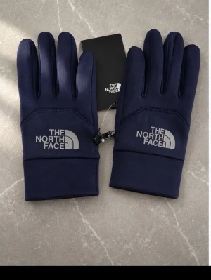 Mörkblå handskar från The North Face - Snygga mörkblå handskar från The North Face med logga på ovansidan. Handskarna har en stilren design och är perfekta för kyliga dagar. Tillverkade i ett mjukt och smidigt material som ger bra komfort och passform.