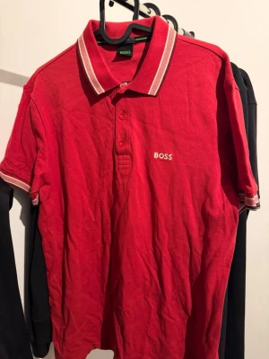 Röd pikétröja från BOSS - Röd pikétröja från BOSS med klassisk krage och knäppning framtill. Tröjan har vita detaljer på kragen och ärmarna samt broderad BOSS-logga på bröstet. Perfekt för en stilren och sportig look.