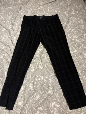 Svarta rutiga kostymbyxor H&M - Snygga svarta kostymbyxor från H&M i slim fit-modell med vitt rutmönster. Byxorna har klassisk passform med pressveck och diskreta slitsar vid benslut. Perfekta för en stilren look.