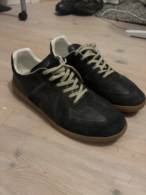 Maison Margiela skor - Snygga svarta sneakers med ovandel i både mocka och slätt läder, vita kontrasterande snören och vit insida. Skorna har en klassisk gummisula i brunt och låg profil som passar till många olika stilar.