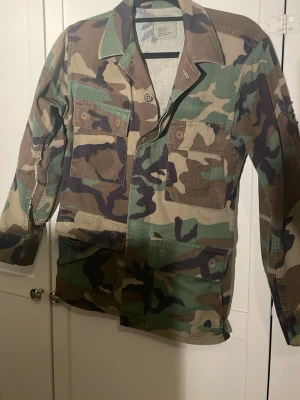 Militärinspirerad camo overshirt - Säljer en overshirt i militärstil med klassiskt camomönster i grönt, brunt och svart. Skjortan har fyra stora fickor framtill, knappar och krage. Tillverkad i slitstark bomull, perfekt för en edgy streetwear-look.