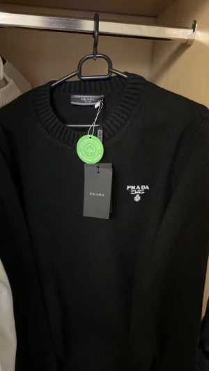 Svart stickad tröja från Prada - Svart stickad tröja från Prada med rund halsringning och diskret logga broderad på bröstet. Tröjan är tillverkad i ekologisk bomull och har en klassisk, stilren design som passar perfekt till många olika outfits.